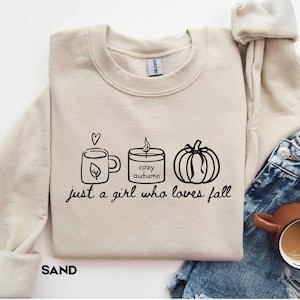 Può includere: Una felpa girocollo color sabbia con una grafica a linee nere che raffigura una tazza con un cuore, una candela con l'etichetta cozy autumn e una zucca. Sotto le icone, il testo recita just a girl who loves fall in carattere corsivo.