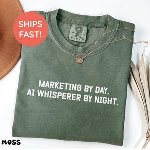 Può includere: Una t-shirt a maniche corte color verde muschio con scollo rotondo. Il capo presenta una scritta bianca stampata sul davanti che recita: "MARKETING BY DAY. AI WHISPERER BY NIGHT." L'indumento ha un'etichetta del marchio Comfort Colors.