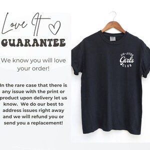 Może przedstawiać: Czarny t-shirt z białym napisem "Jiu-Jitsu Girls Club" z przodu. Koszulka wisi na drewnianym wieszaku. Tekst po lewej stronie obrazu brzmi "Love It Guarantee" i "We know you will love your order!"