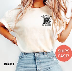 May include: An ivory short-sleeved t-shirt featuring a black graphic on the upper left chest. The graphic depicts a rose surrounded by small stars and the text "DARK ROMANCE BOOK CLUB" printed in a black serif font above and below the rose.