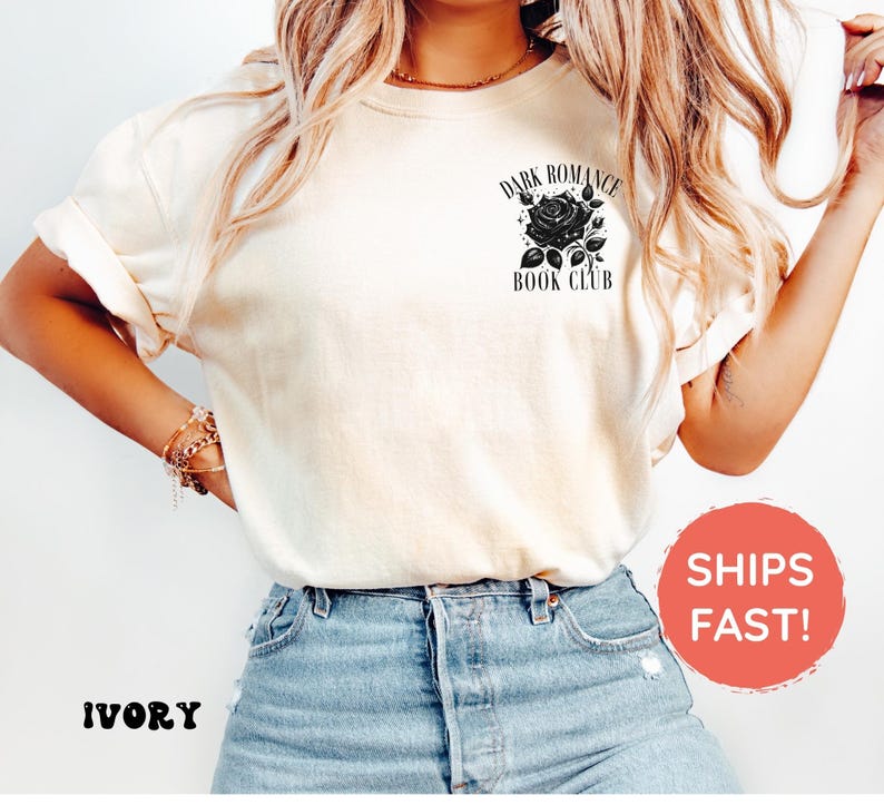 May include: An ivory short-sleeved t-shirt featuring a black graphic on the upper left chest. The graphic depicts a rose surrounded by small stars and the text "DARK ROMANCE BOOK CLUB" printed in a black serif font above and below the rose.