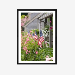 May include: A row of American flags are displayed on a fence in front of a house with pink flowers. The flags are waving in the wind.