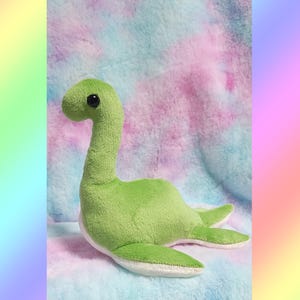 May include: A plush toy shaped like a long-necked dinosaur, featuring a bright green body with a white underside and small black plastic eyes. The soft fabric has a textured appearance, and the creature is depicted in a resting position.