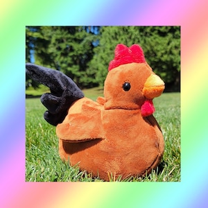 May include: A plush chicken toy featuring a soft brown body, a bright red comb and wattle, a yellow beak, and a dark black tail. The stuffed animal has small black eyes and a rounded, plump shape, crafted from a smooth, velvety fabric material.