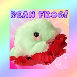 May include: A light green plush frog with black eyes and pink cheeks, resting inside a ruffled red fabric collar. The text BEAN FROG! is printed in blue letters with a white outline at the top of the image against a pastel rainbow background.