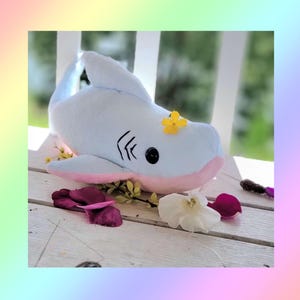 May include: A light blue plush shark toy with a soft pink underside, black embroidered gill slits, and a single black eye. A small yellow flower rests on its head, and the shark is surrounded by scattered white and dark pink flower petals.