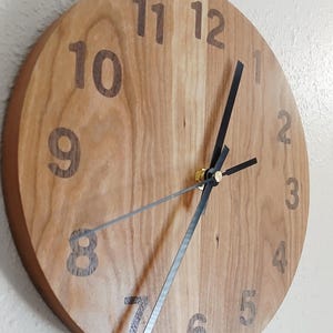 May include: A round wooden clock with a light brown face and dark brown numerals. The clock hands are black, and the second hand is silver. The clock is approximately 12 inches in diameter.