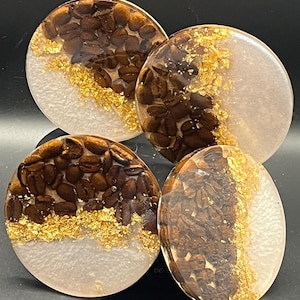 May include: Set of four round coasters. Each coaster features a unique design with embedded coffee beans, gold leaf accents, and a white resin base. The coasters are a decorative and functional accessory.