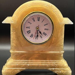 Peut inclure: Une horloge à quartz décorative avec un cadre doré en forme d'arche. Le cadran de l'horloge est rose clair avec des chiffres romains noirs et des aiguilles dorées. La base est rectangulaire et la bordure est dorée.