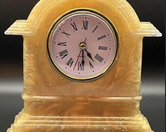 Könnte beinhalten: Eine dekorative Quarzuhr mit einem goldfarbenen, bogenförmigen Rahmen. Das Zifferblatt ist hellrosa mit schwarzen römischen Ziffern und goldenen Zeigern. Die Uhr hat einen rechteckigen Sockel und einen goldfarbenen Rand.