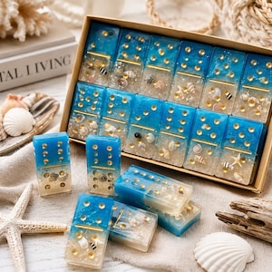 May include: A set of rectangular dominoes crafted from resin, featuring a gradient design that transitions from a deep blue top to a sandy, translucent bottom. Each piece is embedded with miniature seashells and gold-colored pips.