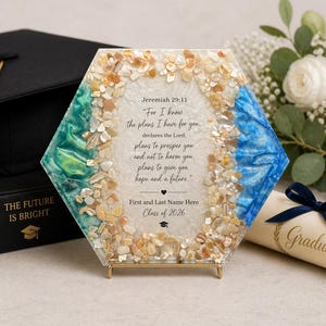 Puede incluir: Un recuerdo de graduación hexagonal con un soporte dorado. La obra de arte presenta una cita de Jeremías 29:11, rodeada de acentos beige y azules. El texto incluye el nombre del graduado y el año de la clase.