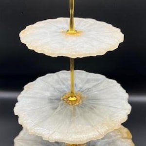 Op de afbeelding: Een serveerstandaard met drie niveaus met een goudkleurige handgreep en steun. De drie niveaus zijn rond, met een wit en grijs marmerpatroon. De standaard is ideaal voor het presenteren van desserts of hapjes op een feest of evenement.