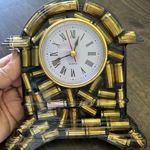 Puede incluir: Un reloj de cuarzo con esfera blanca, manecillas doradas y números romanos. El reloj está encerrado en una resina transparente llena de casquillos dorados. El reloj tiene una base negra y un bisel dorado.