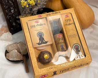 May include: A gift set with the text "Yog Chakra" includes incense, a spray bottle, and a wooden incense holder. The box is tan with a floral design and a clear window. The products feature a meditating figure and colorful chakra symbols.