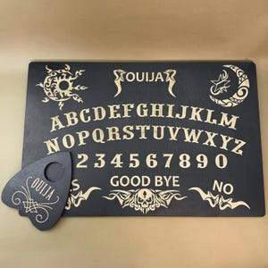 May include: A black wooden Ouija board with gold lettering and symbols. The board features the alphabet, numbers 1-9 and 0, the words "yes", "no", "good bye", and a planchette.