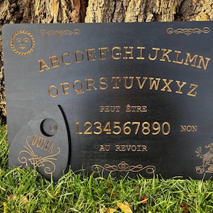 以下が含まれることがあります： 金色の文字とプランシェットが付いた黒いウィジャボード。ボードにはアルファベット、数字、「Peut Être」や「Au Revoir」などのフレーズが記載されています。装飾的な太陽と花の模様が木にエッチングされています。