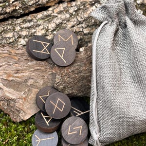 May include: A set of wooden rune stones with gold-colored etched symbols, scattered on a bed of green moss and a piece of wood. A gray drawstring bag is visible on the right side of the image. The runes are dark brown and circular.