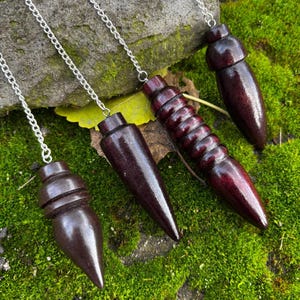 May include: Four dark brown wooden pendulums with silver chains rest on a bed of green moss. Each pendulum has a unique shape, including a conical, cylindrical, and acorn-like design. The wood has a rich, polished finish.