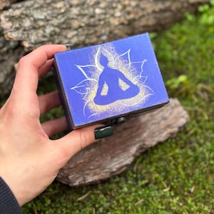 May include: A small, rectangular blue box with a hinged lid. The top features a white and gold illustration of a person in a seated yoga pose. The box is held in a hand against a natural background.