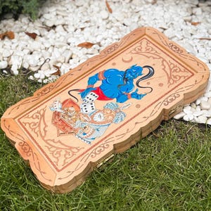 May include: Wooden backgammon board with a detailed illustration of a blue genie, dice, and other figures. The board has a light brown finish with decorative carvings around the edges. The game is set on a bed of green grass and white stones.