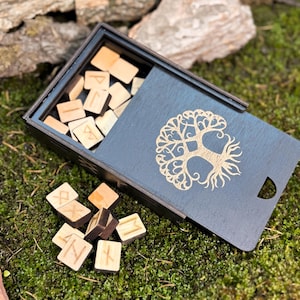 Könnte beinhalten: Eine Holzkiste mit Schiebedeckel, die kleine Runensteine aus Holz enthält. Der Deckel ist mit einem Lebensbaum-Design verziert. Die Kiste und die Steine liegen auf grünem Moos.