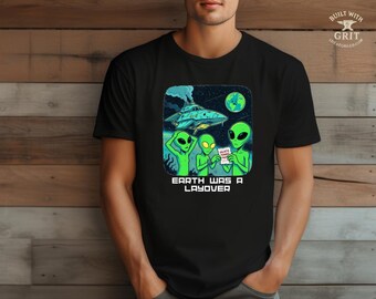 May include: Black t-shirt featuring a cartoon graphic of three green aliens, a spaceship, and the Earth. The aliens are holding a paper that says "Galactic Travel Info". The text "Earth Was A Layover" is printed below the graphic.