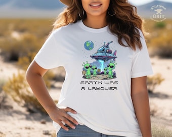 May include: White t-shirt featuring a colorful graphic of a spaceship, aliens, and the Earth. The text "EARTH WAS A LAYOVER" is printed below the image. The design includes green aliens and a blue and pink spaceship.