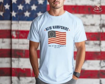 May include: Light blue t-shirt featuring a distressed graphic of the New Hampshire flag with the text "NEW HAMPSHIRE" and "One of the Original 13 Est 1776". The shirt has short sleeves and a crew neck.