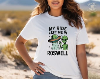 May include: White t-shirt featuring a graphic of a green alien and a UFO with the text "MY RIDE LEFT ME IN ROSWELL". The design is in black, green, and orange. The shirt is a casual wear item.