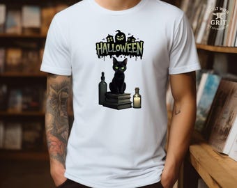 May include: White t-shirt featuring a Halloween design. The graphic includes a black cat with green eyes, sitting on books, with a bottle, a candle, and the word "HALLOWEEN" in green and black. A spooky house and pumpkin are also included.