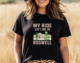 May include: Black t-shirt with the text "MY RIDE LEFT ME IN ROSWELL". The graphic features a green alien, cacti, and desert landscape. The shirt is a crew neck and appears to be made of a soft material.