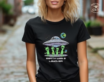May include: Black t-shirt featuring a graphic of a UFO with three green aliens and the Earth. The text "Earth Was a Layover" is printed below the image. The design is centered on the shirt.