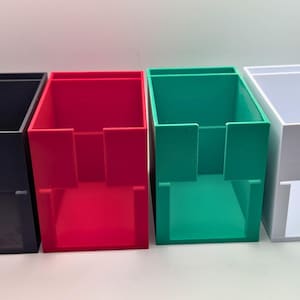 May include: Four rectangular storage bins in black, red, green, and white. Each bin has a rectangular opening and a slot cut out of the front. The bins are made of a solid material and are arranged side by side.