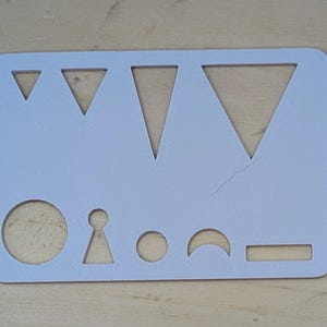 May include: A light blue stencil with various cut-out shapes, including triangles, circles, a keyhole, a crescent, and a rectangle. The stencil is made of a rigid material and is designed for drawing or crafting.