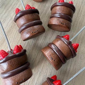 May include: Several miniature chocolate cake ornaments with red strawberry toppings. Each cake has a dark brown frosting and a smiling face. The cakes are attached to silver wires, suggesting they are hanging decorations.