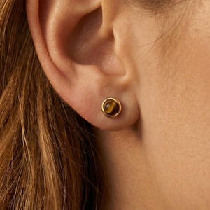 Puede incluir: Primer plano de una oreja con un pendiente de botón dorado. El pendiente presenta una pequeña piedra redonda de ojo de tigre engastada en un bisel dorado. Diseño sencillo y elegante.