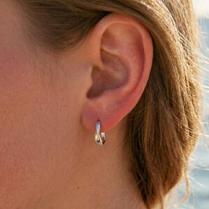 May include: A close-up of a person's ear wearing a small, silver hoop earring. The earring has a twisted design. The background is blurred, with a view of the ocean and sunlight.
