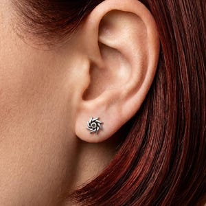 May include: A close-up of a silver stud earring in the shape of a stylized sun or spiral. The earring is worn on an ear, with the focus on the jewelry. The earring has a detailed, textured design.