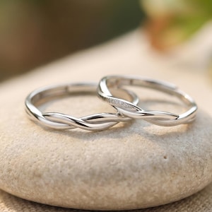 May include: Two silver rings with a twisted design, one with small embedded stones, sit on a smooth, light-coloured stone. The rings are displayed on a neutral-toned fabric, with a blurred green background.