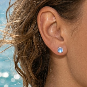 Peut inclure: Gros plan d'une oreille ornée d'une boucle d'oreille ronde bleu clair. La boucle d'oreille a un sertissage argenté et un aspect lumineux et irisé. L'arrière-plan est une vue floue de l'océan.