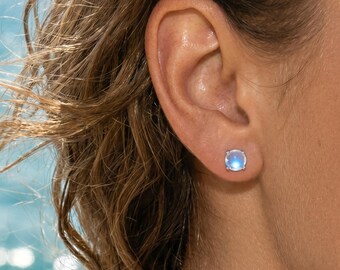 Puede incluir: Primer plano de una oreja con un pendiente de botón redondo azul claro. El pendiente tiene un engaste plateado y un aspecto luminoso e iridiscente. El fondo es una vista borrosa del océano.
