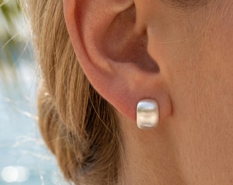 Puede incluir: Primer plano de un pendiente de botón rectangular plateado en una oreja. El pendiente tiene un acabado de metal cepillado y una forma ligeramente redondeada. El fondo está borroso, con toques de azul y verde.