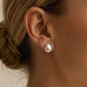 Peut inclure: Une boucle d'oreille à clou unique présentant un design lisse et arrondi en forme de galet avec une finition argentée mate. La boucle d'oreille est positionnée au centre du lobe, affichant une esthétique épurée avec une texture de surface douce.