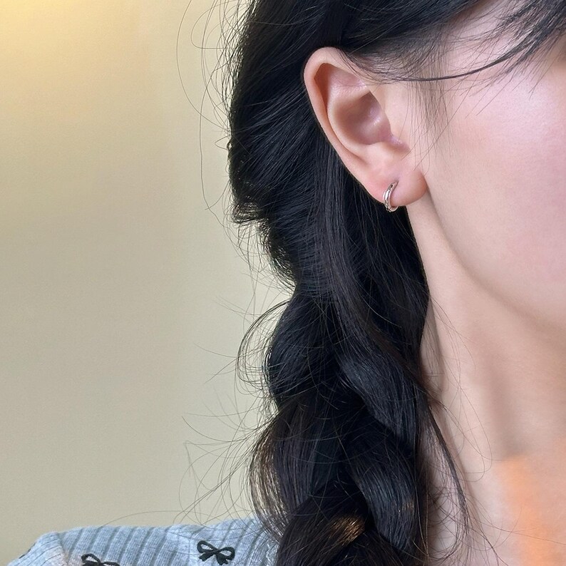 Puede incluir: Primer plano de un pendiente de aro plateado en una oreja. El pendiente es un dise&ntilde;o de aro peque&ntilde;o y sencillo. El pelo oscuro de la persona est&aacute; trenzado y visible en la imagen.