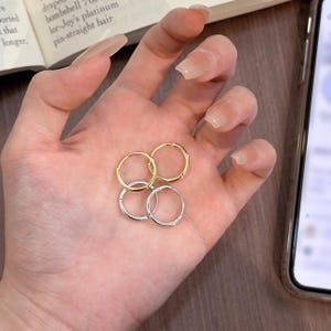 Puede incluir: Cuatro pendientes de aro peque&ntilde;os, dos dorados y dos plateados, se muestran en una mano. Los pendientes tienen un dise&ntilde;o simple y cl&aacute;sico, con forma circular. El fondo es borroso.