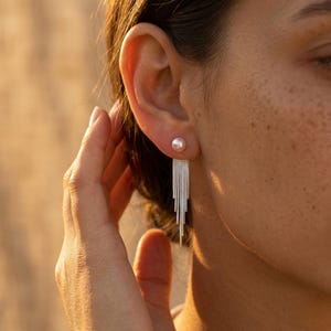 May include: A silver earring featuring a single round pearl stud at the top, from which a series of delicate, tiered silver chains hang downward in a graduated fringe pattern, creating a long, dangling, and layered metallic effect.