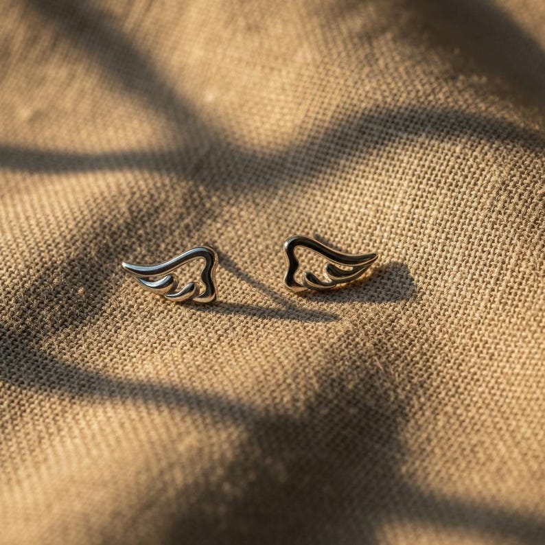 May include: A pair of silver-colored wing-shaped stud earrings. The earrings are small, with a detailed feather design. They are set against a textured, tan background, with soft lighting.