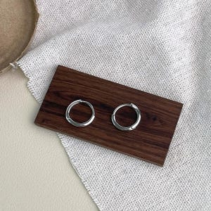 Puede incluir: Un par de pendientes de aro peque&ntilde;os y plateados. Los pendientes se exhiben sobre un rect&aacute;ngulo de madera marr&oacute;n oscuro, sobre una tela texturizada de color neutro. Los aros tienen un dise&ntilde;o simple y minimalista.
