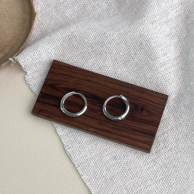 Puede incluir: Un par de pendientes de aro peque&ntilde;os y plateados. Los pendientes se exhiben sobre un rect&aacute;ngulo de madera marr&oacute;n oscuro, sobre una tela texturizada de color neutro. Los aros tienen un dise&ntilde;o simple y minimalista.